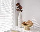 Bulk dried natural badam nut pods in a tall white vase, styled with decorative rattan balls on a gold pedestal bowl.