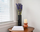 Dark blue dried larkspur flowers in a tall black vase on a wooden table with a candle and stacked white books, natural.