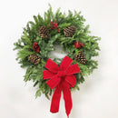 A traditional holiday wreath decorated with green pine branches, brown pinecones, white and red berries, and a red bow in the center.