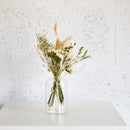 Clear glass bud vase with natural dried flower buds and stems in soft beige and green tones on white surface.