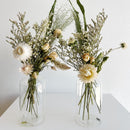 Two clear glass bud vases with natural dried flowers in soft white and beige tones on white surface.