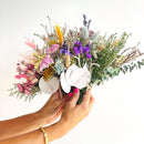 Hands holding small bulk dried flower bundles with pink, purple, yellow, and green natural stems and leaves.