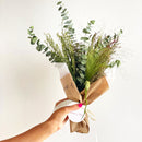 Bouquet of greenery wrapped in brown paper with a white label, held by a hand against a light background.