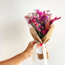 Bouquet of pink and purple flowers wrapped in brown paper held by a hand against a white background