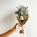 Bouquet of flowers wrapped in brown paper with a white ribbon held by a hand against a light background