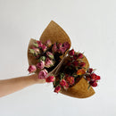 Hand holding two bouquets of dried natural rose sprays in pink and orange tones wrapped in brown paper for wholesale bulk.