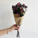 Hand holding a small bouquet of natural dried fuchsia spray roses wrapped in brown kraft paper with twine tie.