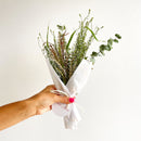 Hand holding a bouquet of greenery wrapped in white paper with a visible brand name on a light background