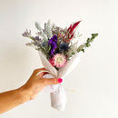 Hand holding small bundle of natural dried flowers in purple, lavender, and pink wrapped in white paper.