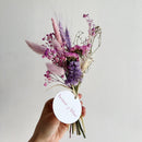 Hand holding a bouquet of purple and pink flowers with a 'house of lilac' tag on a light gray background