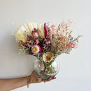 Medium dried flower arrangement in clear round glass vase with natural pink, cream, and green tones, bulk wholesale design.