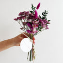 Hand holding a bouquet of purple flowers with a white spherical object against a light background