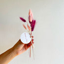 Hand holding a small bouquet of dried pink and purple flowers with a white label against a plain background