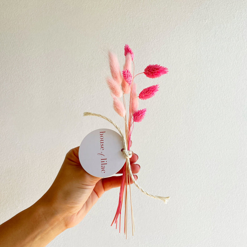 Hand holding a small bouquet of pink flowers with a 'house of lilac' tag against a plain background
