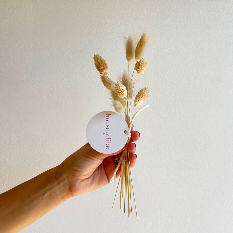 Hand holding a bouquet of dried flowers with a 'house of liac' label against a plain background
