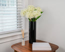 White dried hydrangea spray with natural blooms arranged in a tall black vase on a wooden table, showing wholesale bulk.