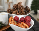 Bulk dried natural pomegranates and orange slices in white bowls with pine cones in background.
