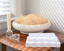 Natural dried wood excelsior in a white bowl, displayed with white boxes and glass candle holders on a wooden table in.