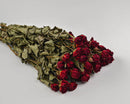 A dried rose bouquet with deep red flowers and green leaves laid out on a white surface.