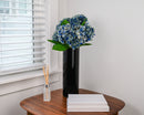 Two-tone blue hydrangea stems arranged in a black vase on a wooden table, natural dried wholesale bulk floral display.