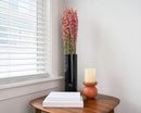 Tall black vase holding natural dried pink larkspur flowers on wooden table with books and candle.