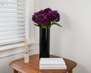 Eggplant dried hydrangea spray with green leaves in tall black vase on wooden table, natural bulk decor.