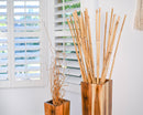 Dried natural river cane bamboo sticks in bulk arranged in tall wooden vase by window blinds.
